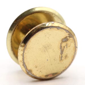 May include: Close-up of a vintage brass cabinet knob. The knob has a circular design with a slightly domed top and a flat, circular base. The brass has a warm, golden hue with some signs of wear.