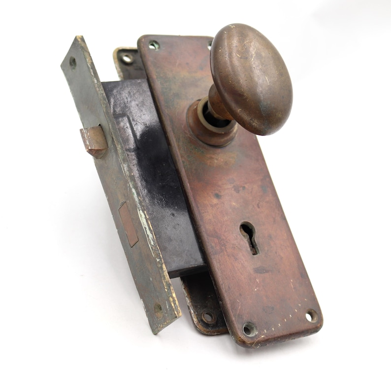 May include: Antique door hardware set with a tarnished bronze finish. The set includes a rectangular faceplate with a keyhole, an oval doorknob, and a latch mechanism. The metal shows signs of age and wear, with a patinaed appearance.