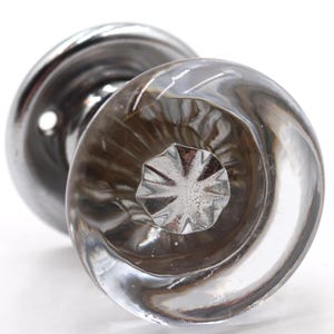 Puede incluir: Un pomo de puerta de cristal transparente con base cromada. El pomo de cristal tiene un diseño en espiral y un patrón de estrella en el centro. Este herraje es probablemente para una puerta o un armario.