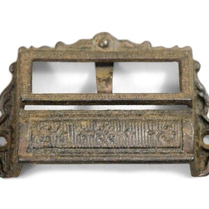 Pode incluir: Puxador de gaveta antigo de ferro fundido com uma abertura retangular e um design ornamentado. O puxador tem um acabamento marrom rústico.