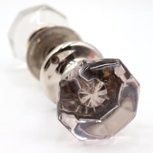 Könnte beinhalten: Ein dekorativer Türknauf mit einem facettierten, rauchgrauen Glasende. Der Knauf hat einen silberfarbenen Metallsockel und ein klares Glasende. Das Design ist vintage und elegant, geeignet für die Inneneinrichtung.