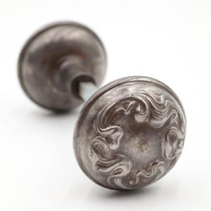 May include: A pair of round, metallic door knobs with a dark, weathered finish. Each knob features a raised, swirling embossed pattern across its surface. The two knobs are connected by a central metal spindle, showing a detailed, textured design.