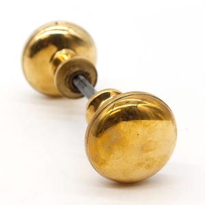 Può includere: Una coppia di pomelli per porte in ottone lucido con un design semplice. I pomelli sono collegati da un'asta di metallo.