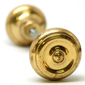 Peut inclure: Deux boutons de porte anciens en laiton doré. Les boutons ont une forme ronde avec un motif circulaire décoratif sur le devant. La quincaillerie est posée sur un fond blanc, soulignant l'aspect vieilli du métal.