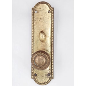 May include: Vintage brass door handle with a rope-style border. The handle has a round knob and a long, rectangular backplate. The brass has a worn, aged look, indicating it is antique. The design is ornate and classic.