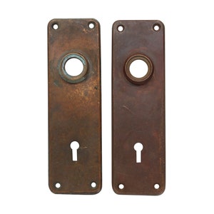 May include: Two antique brass door plates with keyholes and round knob holes. The plates are rectangular and have a worn, aged finish.