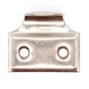 Puede incluir: Gancho de metal plateado con dos agujeros. El gancho tiene una parte superior redondeada y una base rectangular.