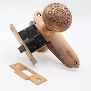 Puede incluir: Juego de herrajes de puerta de bronce antiguo, que incluye un pomo decorativo con un diseño floral en relieve, una placa para la cerradura y una cerradura de embutir. El conjunto es de bronce.