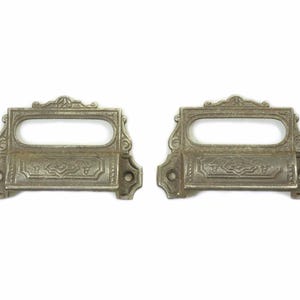 May include: Two antique silver drawer pulls with a rectangular opening for a label. The pulls have an ornate design with a raised edge.