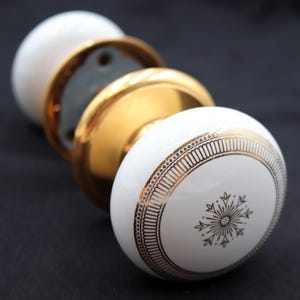 May include: White and gold door knobs with a decorative snowflake design. The knobs have a gold base and a white, rounded top with a gold border. The hardware is set against a dark background.