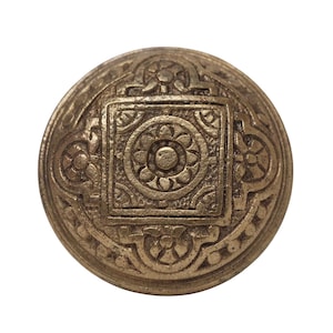 May include: A round, gold-toned metal button with an intricate, embossed design. The centre of the button features a square with a floral pattern.
