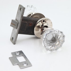 May include: Vintage door hardware set featuring a crystal glass doorknob, a metal latch, and a strike plate. The doorknob has a faceted design. The metal components have a silver finish. Ideal for home restoration projects.