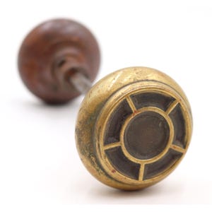 May include: Vintage brass door knob with a circular design and a dark brown wooden knob in the background. The brass knob has a weathered appearance, suggesting age and use. The design features a central circle with radiating lines.