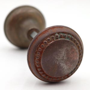 Könnte beinhalten: Antiker Türknauf aus Bronze mit rundem, strukturiertem Design. Der Knauf hat einen dekorativen Rand aus kleinen, erhabenen Kreisen. Das Metall hat ein verwittertes Aussehen mit Anklängen von grüner Oxidation. Auf weißem Hintergrund.