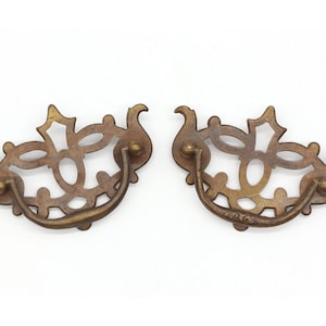 May include: Two ornate brass drawer pulls with a decorative, openwork design. The pulls have a simple, curved handle.
