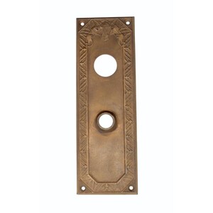 May include: A brass door plate with an ornate design and two keyholes. The plate is rectangular and has a slightly raised edge.