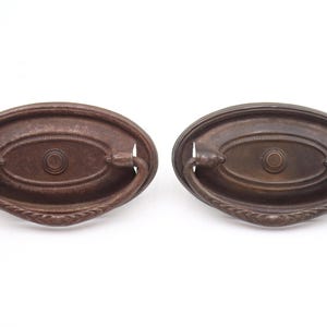 Puede incluir: Dos tiradores de cajón de estilo antiguo con acabado marrón oscuro. Cada tirador tiene una placa trasera ovalada con un detalle circular central y un asa curvada. Los tiradores son de metal.