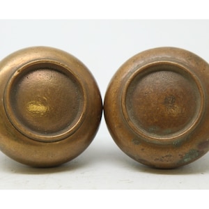 May include: Two antique bronze door knobs. The knobs are round with a slightly flattened top and a circular indentation on the bottom. The bronze has a warm, aged patina, with some areas showing darker oxidation.