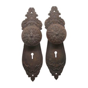 Puede incluir: Dos pomos de puerta antiguos con diseños ornamentados y agujeros de llave. Los pomos son de metal oscuro con un acabado rústico.