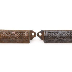 May include: Two antique cast iron drawer pulls with ornate designs. The pull on the left is a rusty brown color, while the pull on the right is a dark gray color.