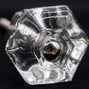 Puede incluir: Un pomo de armario de vidrio transparente y facetado con un tornillo de metal plateado. El pomo tiene forma hexagonal con una hendidura circular central. El vidrio refleja la luz.