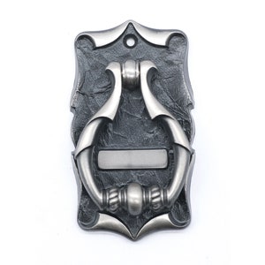 Puede incluir: Un llamador de puerta de plata con un diseño decorativo. El llamador está unido a una placa rectangular con una placa de nombre.