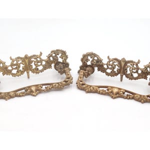 May include: Two antique brass drawer pulls with ornate, scrolling designs. The pulls have a curved handle and decorative floral and leaf accents. The hardware is a warm, aged brass color, suggesting vintage or vintage-inspired furniture.