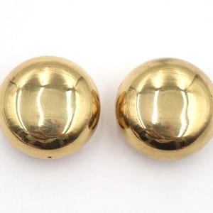 May include: A pair of gold-coloured, spherical clip-on earrings. Each earring has a smooth, reflective surface, catching the light. The earrings are a simple, classic design, suitable for various occasions. The earrings are displayed on a white background.