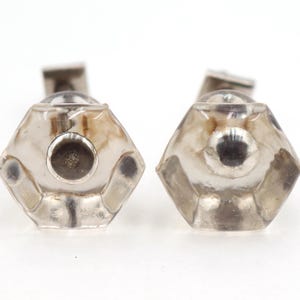 Puede incluir: Dos pomos de armario de cristal transparente facetado con herrajes de metal plateado. Los pomos tienen una forma geométrica hexagonal con un detalle circular central. Los herrajes son rectangulares.