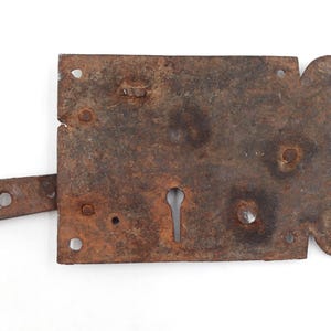 May include: A rusty, antique metal door latch with a keyhole and a sliding bolt. The latch is rectangular with rounded edges and several rivet holes. The metal has a weathered, aged appearance.