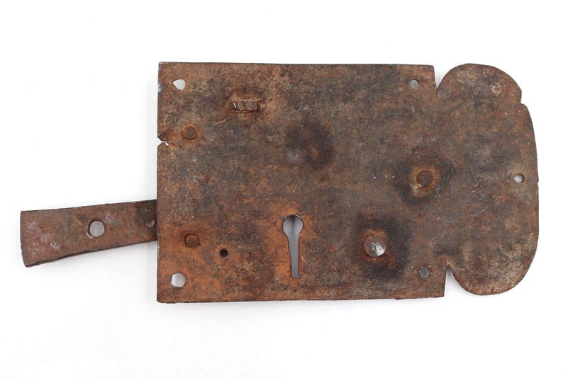 May include: A rusty, antique metal door latch with a keyhole and a sliding bolt. The latch is rectangular with rounded edges and several rivet holes. The metal has a weathered, aged appearance.