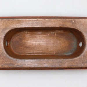 Pode incluir: Um puxador de gaveta retangular, de estilo antigo, com uma pega oval rebaixada. O puxador é feito de um metal com tom de cobre com um acabamento envelhecido, mostrando variações de cor e textura. Dois pequenos orifícios são visíveis em cada extremidade do oval.