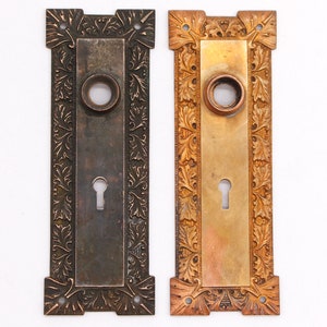 Op de afbeelding: Twee antieke deurplaten met sierlijke bloemmotieven. De ene plaat is in een donker messingkleurige afwerking en de andere in een gepolijste messingkleurige afwerking.
