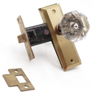 Puede incluir: Un juego de pomo de puerta antiguo con un pomo de cristal transparente facetado, una placa trasera de latón y una placa de cierre a juego. El juego incluye un mecanismo de cierre de metal negro, que muestra una estética vintage.