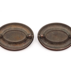 May include: Two antique bronze oval drawer pulls. Each pull has a central oval panel, a beaded border, and small round knobs on each end. The pulls are set against a white background.