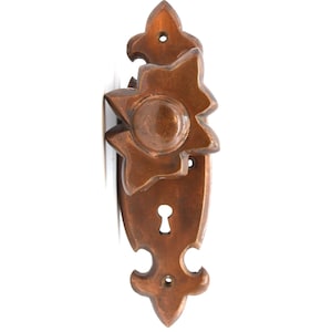 Peut inclure: Poignée de porte en bronze orné avec un motif en étoile sur le bouton. La poignée comporte un trou de serrure et des ornements décoratifs. La quincaillerie est montée sur une surface blanche.