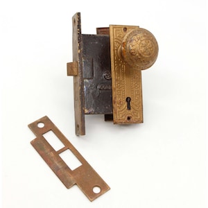 Może przedstawiać: Antyczny zestaw zamka i klamki do drzwi z mosiądzu z ozdobnym wzorem. Zamek ma dziurkę od klucza, a klamka ma okrągły kształt z wypukłym wzorem. Zestaw zawiera płytkę przylgową.