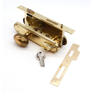 以下が含まれることがあります： 真鍮製のドアロックセット。楕円形のドアノブ、鍵、長方形のストライクプレートが含まれています。 ロック機構が見え、複雑なデザインが示されています。 ハードウェアはヴィンテージ感があります。