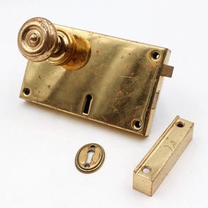 May include: A vintage brass door lock set, comprising a rectangular plate with a keyhole, a round doorknob, and a separate strike plate. An oval escutcheon is also included. The hardware has a warm, aged patina.