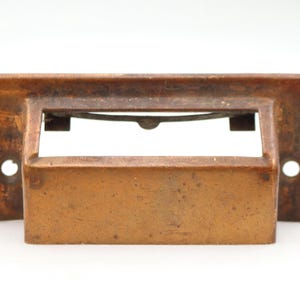 May include: A rectangular, weathered copper-toned metal drawer pull featuring a central label holder slot. The hardware has a flat mounting plate with two circular screw holes on the sides and a protruding handle section with a visible metal spring.