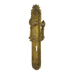 May include: A vintage brass door handle with intricate floral and geometric designs. The handle has a keyhole and two ornate knobs.