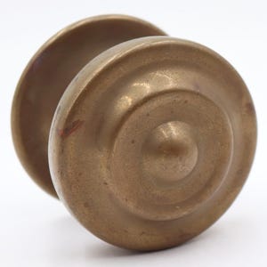 May include: A vintage brass door knob with a circular design. The knob has a warm, golden-brown color and a slightly aged appearance. The design features concentric circles and a central, rounded protrusion.