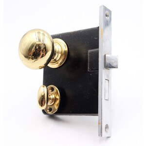 May include: A vintage door lock with a brass doorknob and a polished chrome strike plate. The lock is mounted on a black rectangular plate. The doorknob is round and shiny, and the strike plate is rectangular with screw holes.