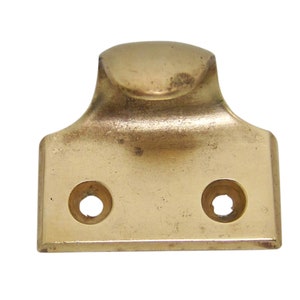 May include: A brass window sash lift with two screw holes. The lift has a rounded top and a rectangular base.