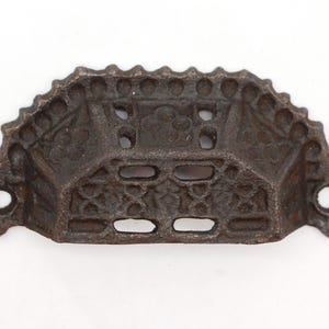 May include: An antique-style drawer pull with a dark brown, aged finish. The cast iron pull features an ornate, floral design and two mounting holes. The handle has a scalloped top edge and a rectangular shape. Suitable for furniture restoration.