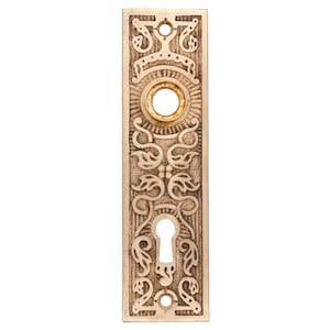 May include: A vintage brass door plate with an ornate design featuring a keyhole and a round hole. The plate has a worn, antique finish.