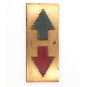 May include: A rectangular brass elevator button panel with an upward-pointing green arrow and a downward-pointing red arrow. The panel has two small circular holes and a gold finish.