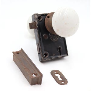 May include: An antique door knob set with white porcelain knobs, a black metal plate, and a separate rusty metal strike plate and keyhole cover. The hardware is likely from the early 20th century.