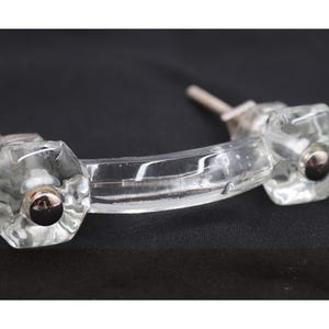 May include: Two clear glass cabinet pulls with a faceted, hexagonal design and silver-colored hardware. The pulls have a curved, rectangular connecting bar. The hardware is silver-colored. The pulls are on a black background.
