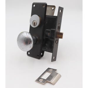 May include: A vintage door lock set with a black metal plate, a silver doorknob, and a keyhole. The set includes a separate silver strike plate. The lock is likely for a traditional or antique door.